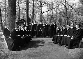 About 24 Christian Brothers dressed in their traditional black robes, white rabats and skull caps are gathered at a clearing in the woods.  Half of them sit on two benches facing each other and the other half stand facing the camera.   In the background there is a cabin with a cross on it.